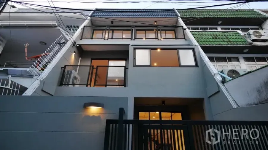 Townhouse for rent in Watthana, Bangkok - Three-storey grey townhouse facade with balconies and a black gate lit at dusk.