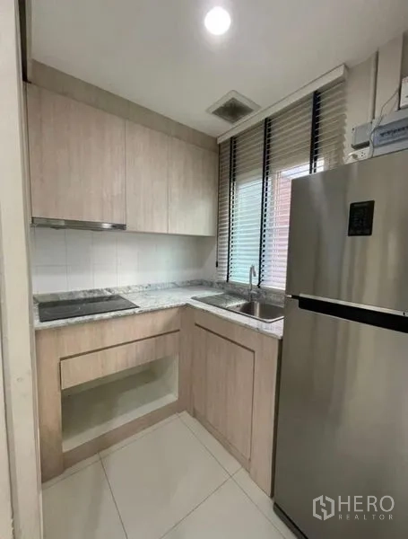 Townhouse for sale - Compact L-shaped kitchen with light wood cabinets, stone countertop, sink, cooktop, and stainless refrigerator.