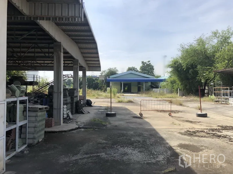 Warehouse for rent or sale in Lam Luk Ka, Pathum Thani - Open-sided storage shed with metal racks and a wide yard leading to a blue-roof building.