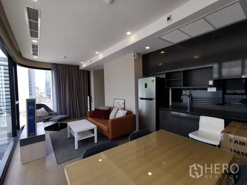 Condominium for rent or sale in Watthana, Bangkok - Open‑plan living and dining area beside full‑height windows, with a sleek black kitchen and stainless refrigerator.