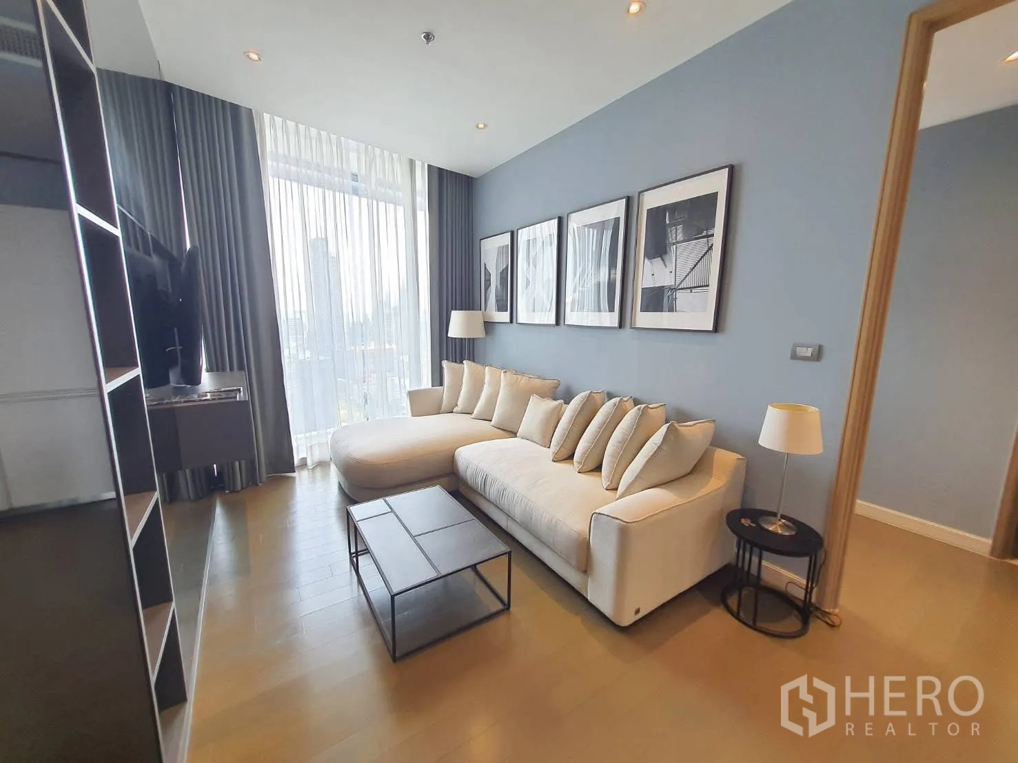 公寓 for rent in Pathum Wan, Bangkok - 现代客厅，长形米色沙发、墙面装饰画，落地窗配纱帘，可观城市景观。