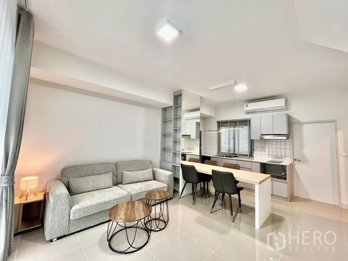 Townhouse for rent in Bang Phli, Samut Prakan - Bright open-plan living room with grey sofa, nesting coffee tables, and a modern kitchen with breakfast island.
