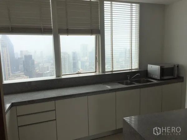 Condominium for rent in Khlong Toei, Bangkok - Windowed kitchen with long granite countertops, double sink, microwave and city views through large blinds-covered windows.