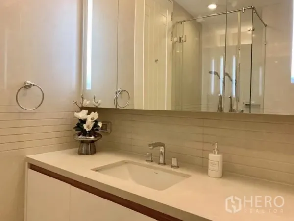 Condominium for rent in Pathum Wan, Bangkok - Modern bathroom with long vanity, under-mount sink, mirrored cabinets, chrome fixtures, and glass-enclosed shower in neutral tones.