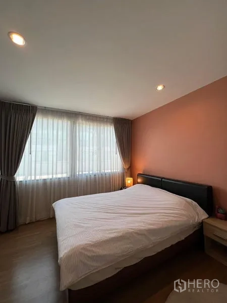 Townhouse for rent in Saphan Sung, Bangkok - Bright bedroom with large curtained windows, peach accent wall and a queen bed with bedside lamp.