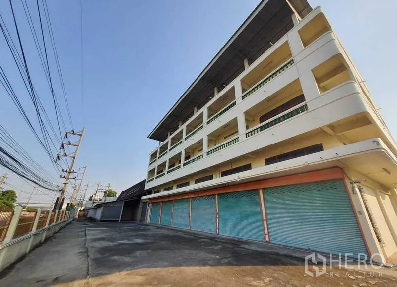 Warehouse for rent or sale in Mueang Pathum Thani, Pathum Thani - Four-storey office and shopfront building with blue roller shutters beside the warehouse complex.