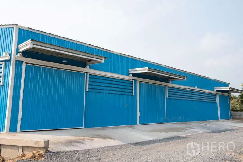 Warehouse for rent in Khlong Luang, Pathum Thani - Long blue warehouse frontage with multiple canopies and a wide concrete ramp for truck loading.