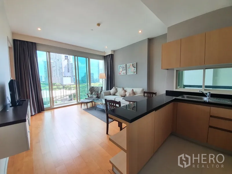 Condominium for rent in Chatuchak, Bangkok - Open-plan space with U-shaped kitchen, black countertop and wide windows framing city and park views.