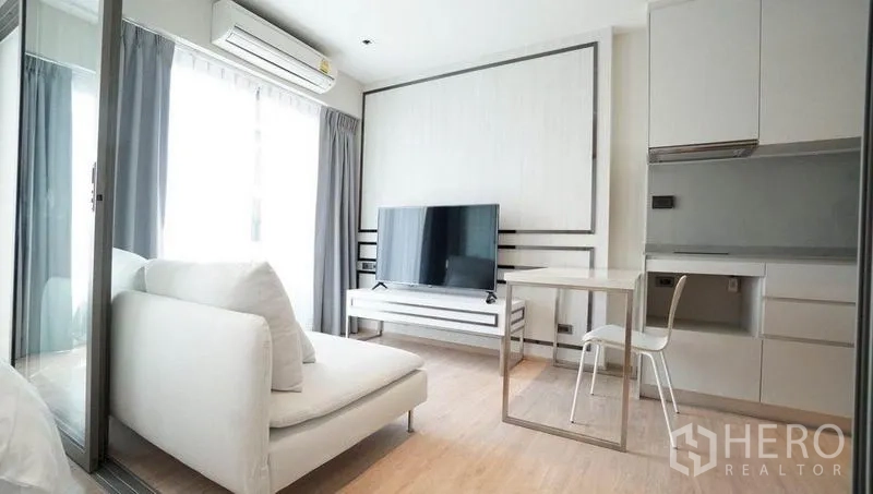 Condominium for sale in Khlong Toei, Bangkok - Light-filled living room with white sofa, large TV, dining table for two and compact kitchenette.