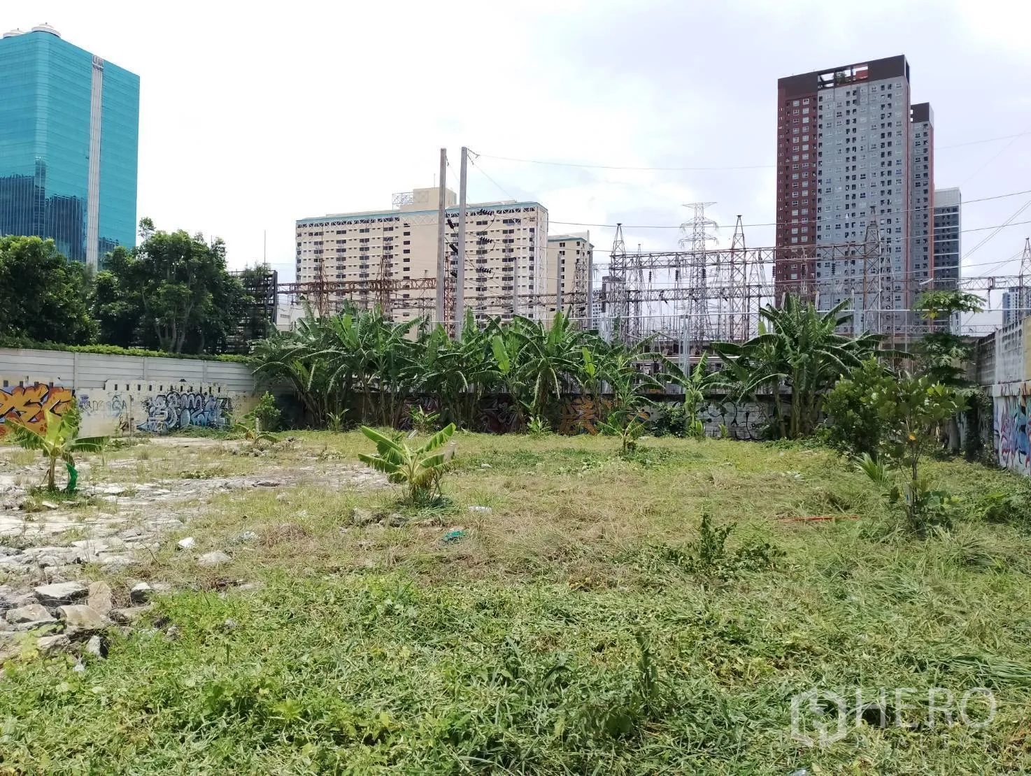 Land for sale in Suan Luang, Bangkok - Open plot with light vegetation and surrounding walls, with mid‑ and high‑rise buildings beyond.