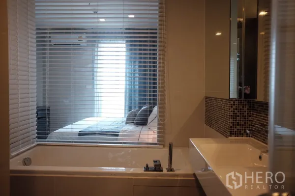 Condominium for rent or sale in Khlong Toei, Bangkok - Modern bathroom with soaking tub, vanity and a glass window with blinds looking toward the bedroom.