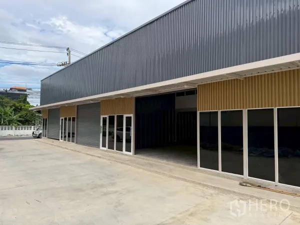 Warehouse for rent in Suan Luang, Bangkok - Angled view of modern warehouse units with dark corrugated facade, multiple glass entrances and a large driveway for trucks.