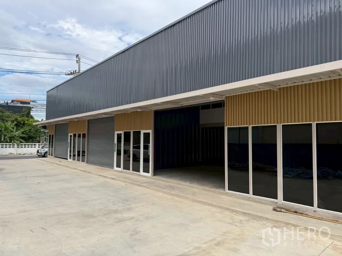 Warehouse for rent in Suan Luang, Bangkok - Angled view of modern warehouse units with dark corrugated facade, multiple glass entrances and a large driveway for trucks.