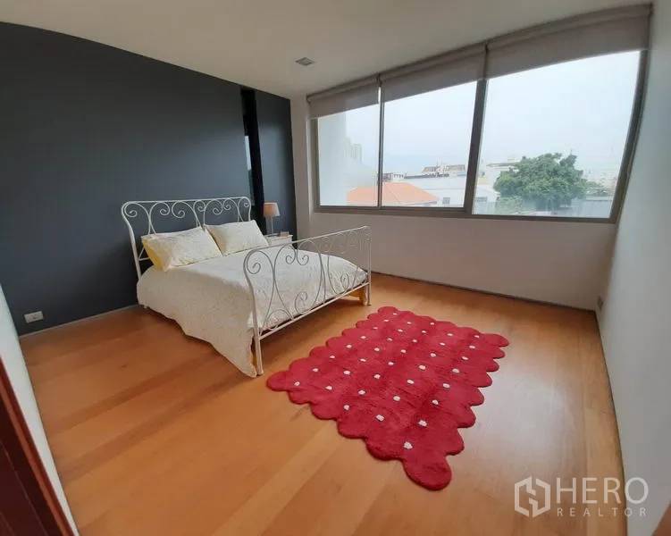 Condominium for rent in Khlong Toei, Bangkok - Bright second bedroom with large window, metal-frame bed and red rug on wood floors.