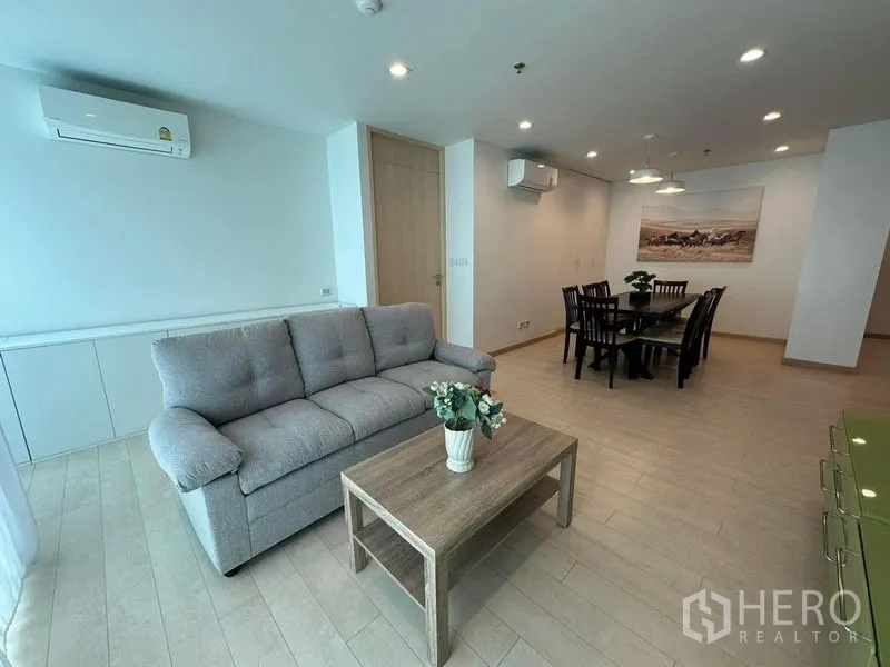 Condominium for rent or sale - Bright open-plan living and dining area with a grey sofa, wood floors, recessed lighting and wall-mounted air conditioners.