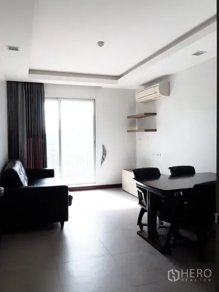 Condominium for rent in Huai Khwang, Bangkok - Bright living and dining area with sliding balcony door, black sofa, table, and wall-mounted air conditioner.
