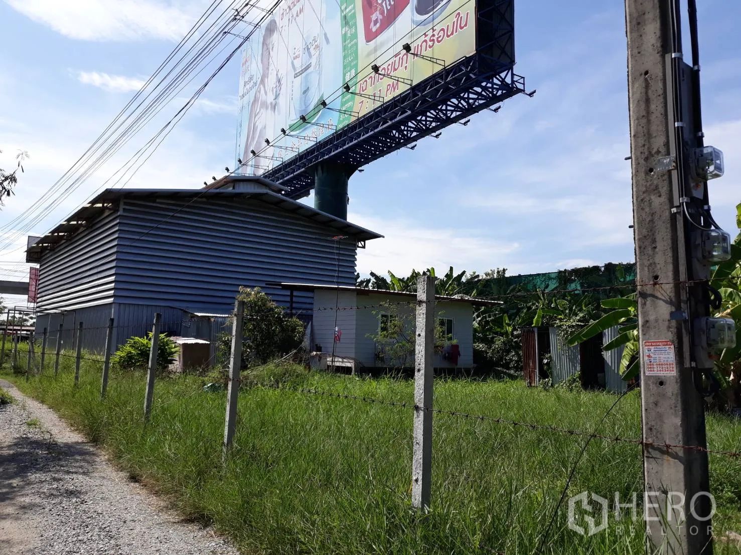 公寓 出租在Pathum Wan, Bangkok - 金属夹芯建筑与小屋的侧面，带铁丝网围栏，旁边有大型路边广告牌。