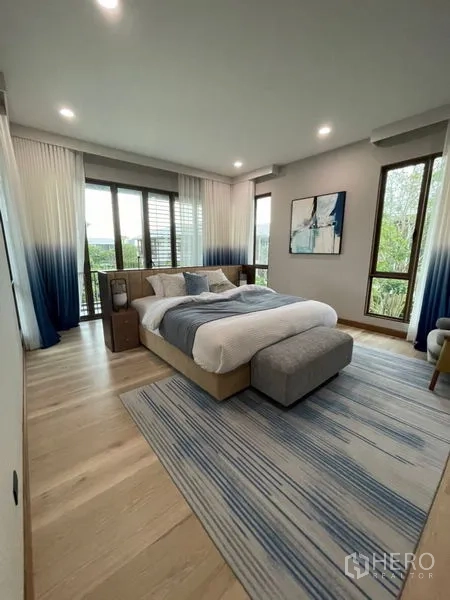 Detached House for sale in Bang Kapi, Bangkok - Bedroom with large windows, blue curtains, area rug and upholstered bed facing greenery.