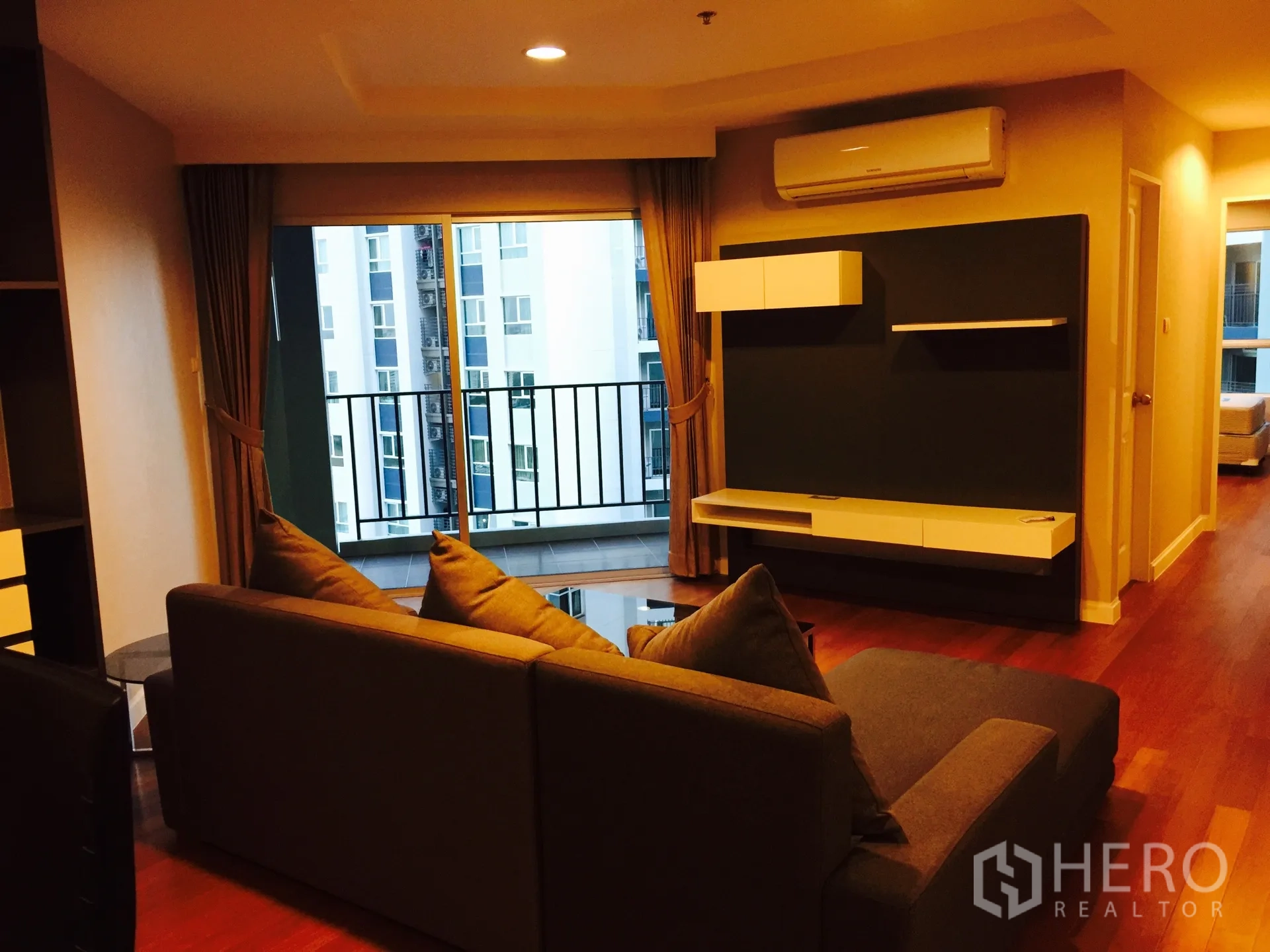 คอนโด ให้เช่าในHuai Khwang, Bangkok - ห้องนั่งเล่นอบอุ่น โซฟาตัวแอล พื้นไม้ และระเบียงมองเห็นอาคารเมือง