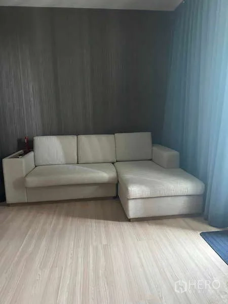 Condominium for sale in Phaya Thai, Bangkok - Beige L‑shaped sofa against a textured accent wall on wood flooring.