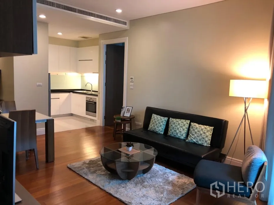 Condominium for rent or sale in Khlong Toei, Bangkok - Open-plan living area with leather sofa, round glass coffee table, wood floors, and view to a white L-shaped kitchen.