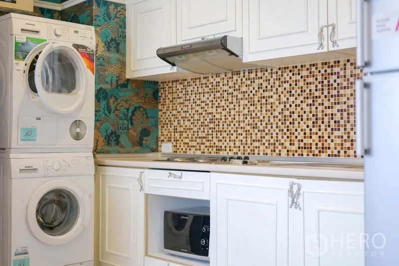 Condominium for rent in Khlong Toei, Bangkok - Compact kitchen with white cabinets, mosaic backsplash, cooktop, microwave, and stacked washer-dryer.