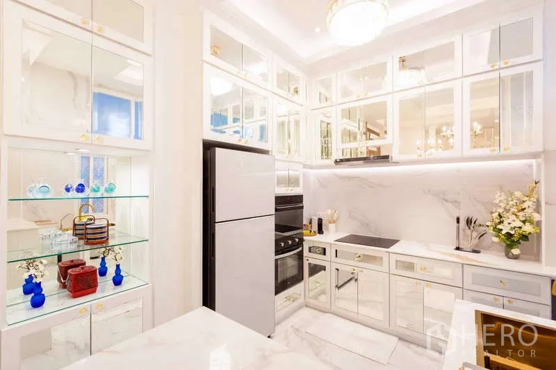 Townhouse for rent in Lat Phrao, Bangkok - Modern white kitchen with mirrored cabinets, built-in oven, cooktop and glass display shelving.