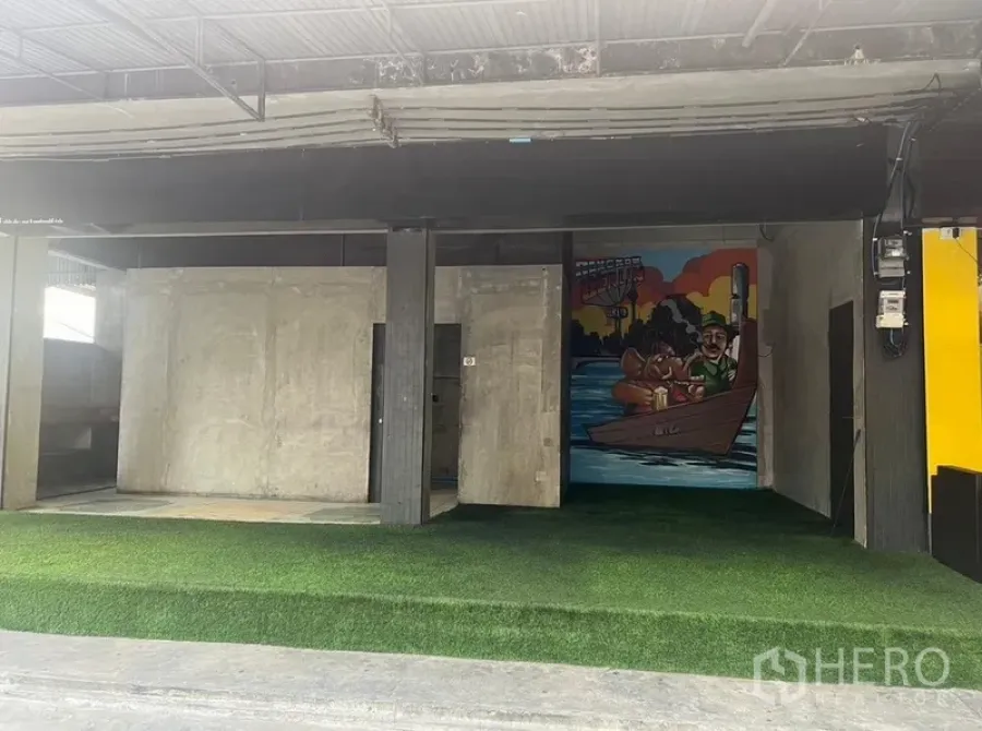 Retail Space for rent in Watthana, Bangkok - Open-front retail bay with concrete walls, artificial grass flooring, and a colorful mural beneath a metal awning.