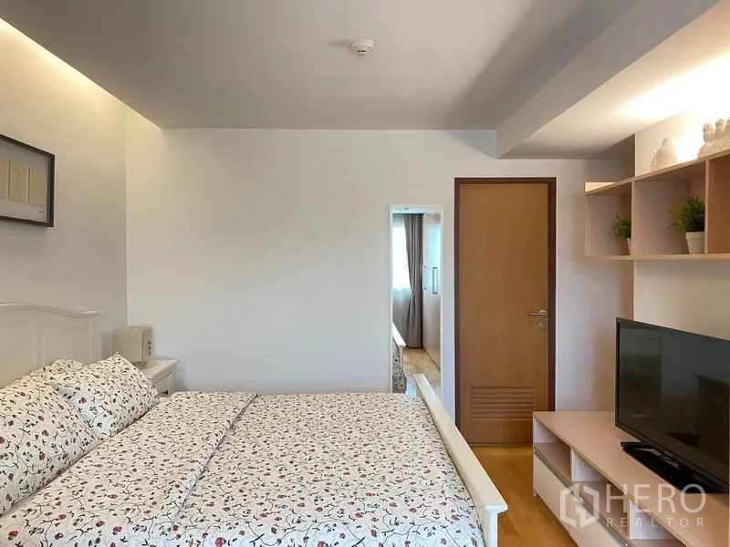 公寓 出租在Phra Khanong, Bangkok - 温馨卧室，电视柜与墙面置物架，对面为花卉床品。