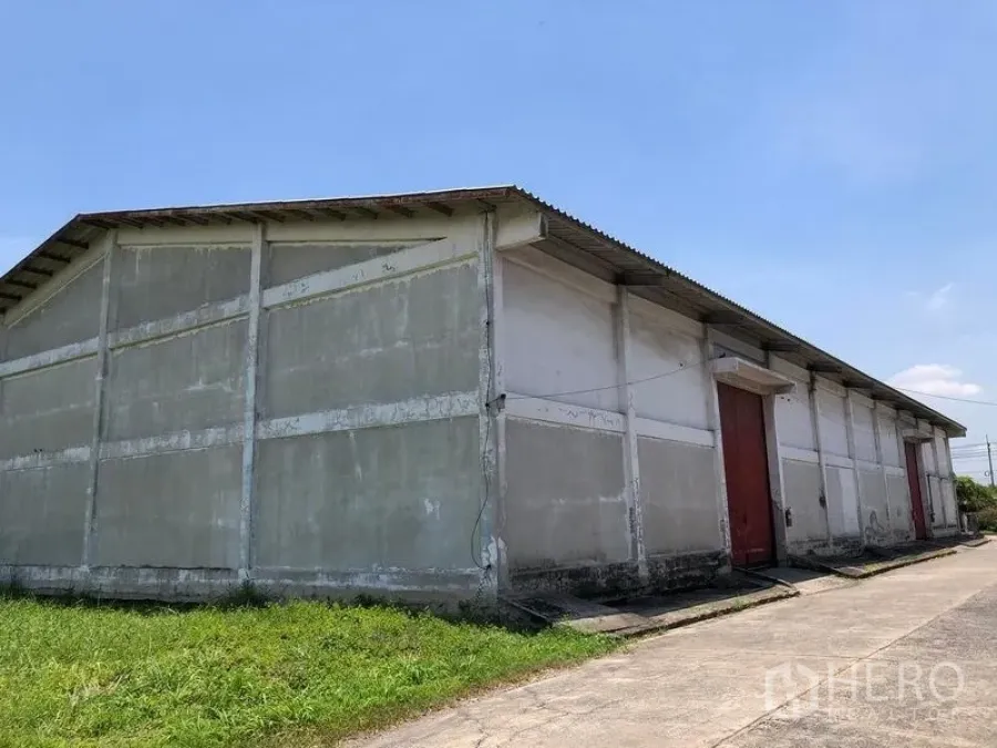 货栈 出租在Lat Krabang, Bangkok - 仓库外观，单层混凝土结构，红色滑动大门、金属坡屋顶与混凝土车道，晴空下。