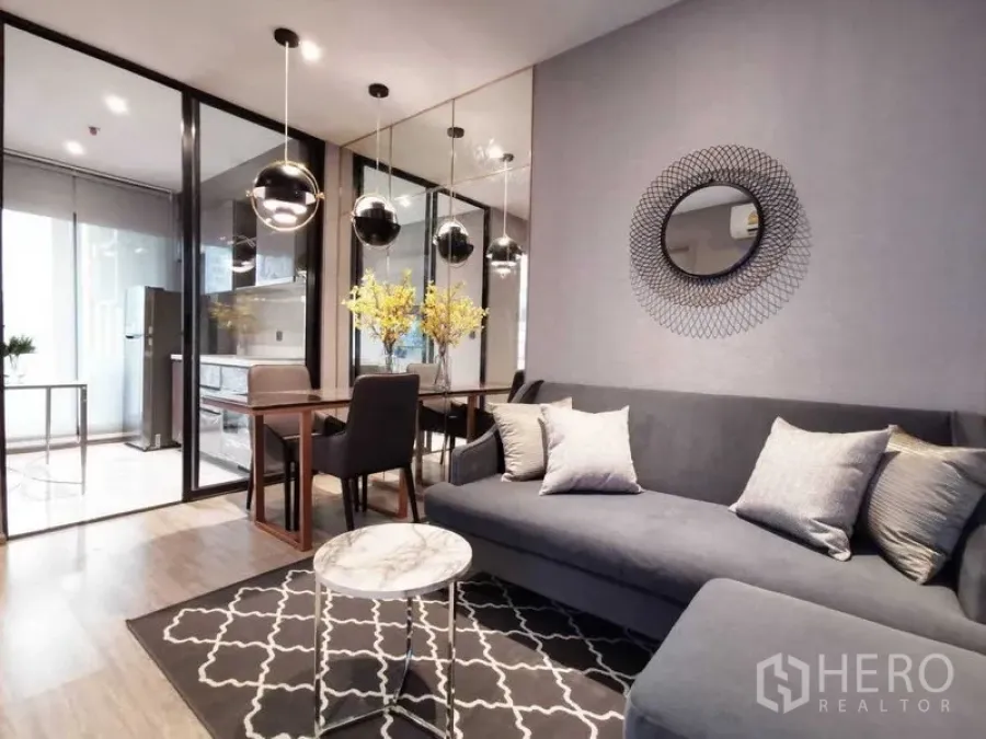 Condominium for sale in Watthana, Bangkok - Living and dining space featuring a grey sofa, round mirror and mirrored wall, marble coffee table, and glass doors to the kitchen.