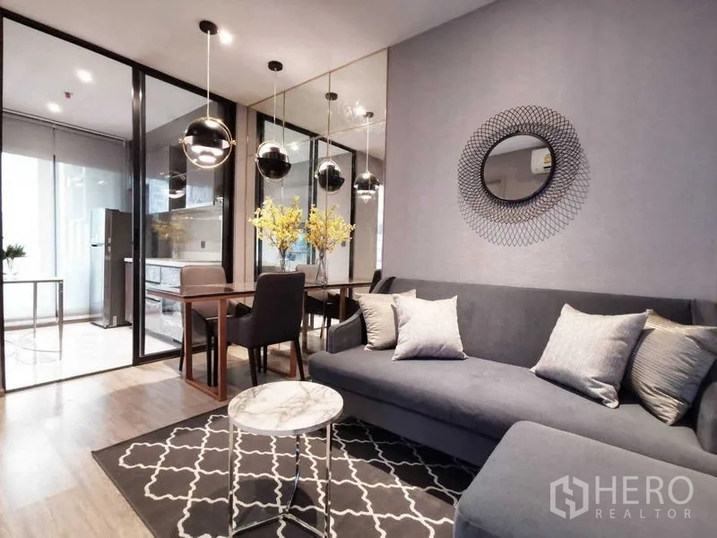 Condominium for sale in Watthana, Bangkok - Living and dining space featuring a grey sofa, round mirror and mirrored wall, marble coffee table, and glass doors to the kitchen.