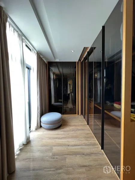 Townhouse for rent in Saphan Sung, Bangkok - Walk-in closet corridor with glass wardrobes, natural light, and round ottoman.