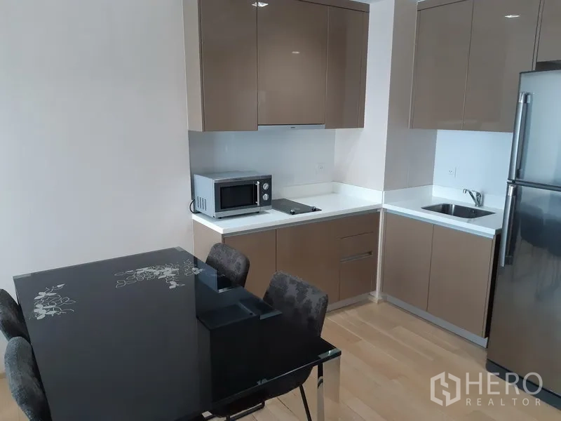 Condominium for rent in Khlong Toei, Bangkok - Modern condo kitchen with glossy taupe cabinets, microwave, cooktop, sink, fridge, and a black glass dining table with four chairs.