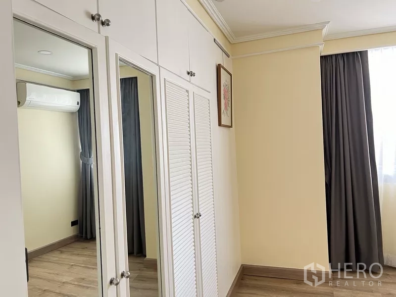 Condominium for rent in Khlong Toei, Bangkok - Second bedroom with white built-in wardrobes, mirrored panels and air conditioner.