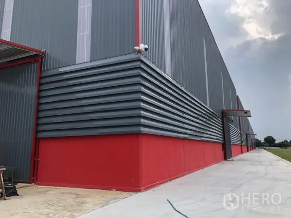 货栈 出租在Khlong Luang, Pathum Thani - 仓库转角立面，波纹金属板与红色基座，并设CCTV与带雨棚的入口。