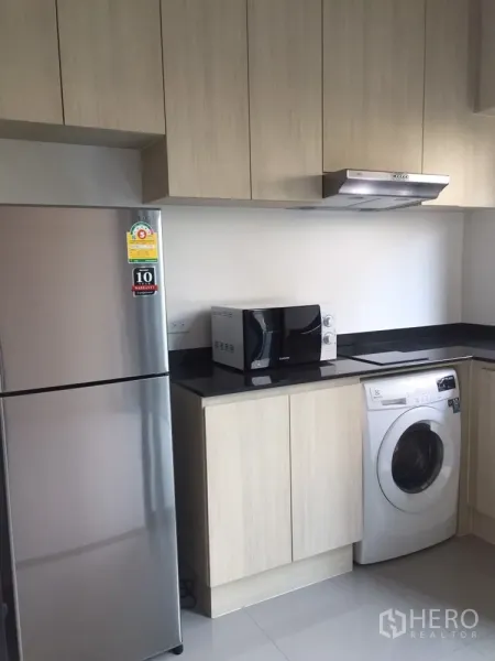Townhouse for rent in Suan Luang, Bangkok - Modern kitchen with light wood cabinets, black countertops, refrigerator, microwave, and washing machine.