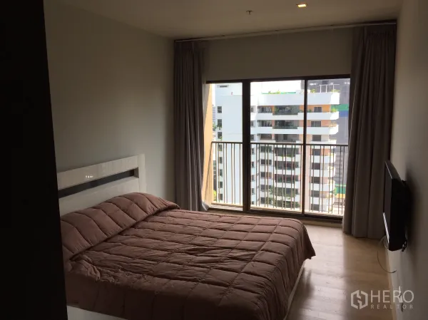 公寓 出租在Bangkok - 明亮卧室配落地窗与城市景观阳台，浅色木地板与暖色床品。