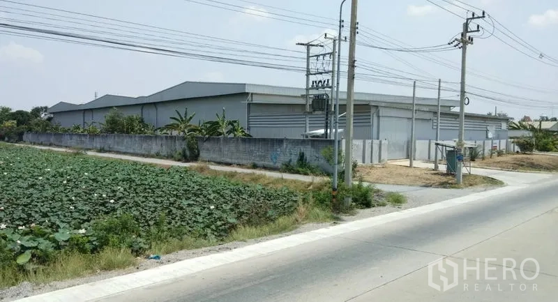 Warehouse for rent in Pathum Thani - Gated warehouse compound beside a main road with perimeter wall and power lines.