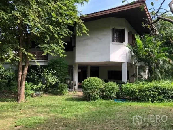 Detached House for sale in Bang Na, Bangkok - Two-storey house with a covered terrace and landscaped lawn beneath shady trees.