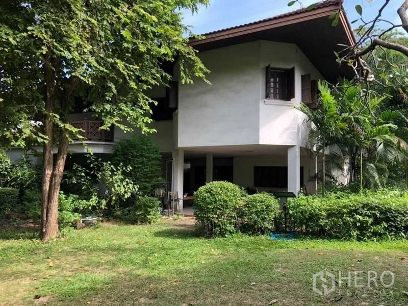 Detached House for sale in Bang Na, Bangkok - Two-storey house with a covered terrace and landscaped lawn beneath shady trees.