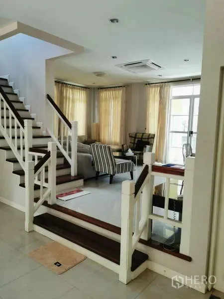 Detached House for rent - Open-plan living area with a white staircase, armchairs, and large windows dressed with beige curtains.