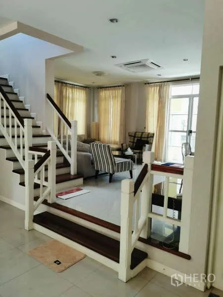 Detached House for rent - Open-plan living area with a white staircase, armchairs, and large windows dressed with beige curtains.