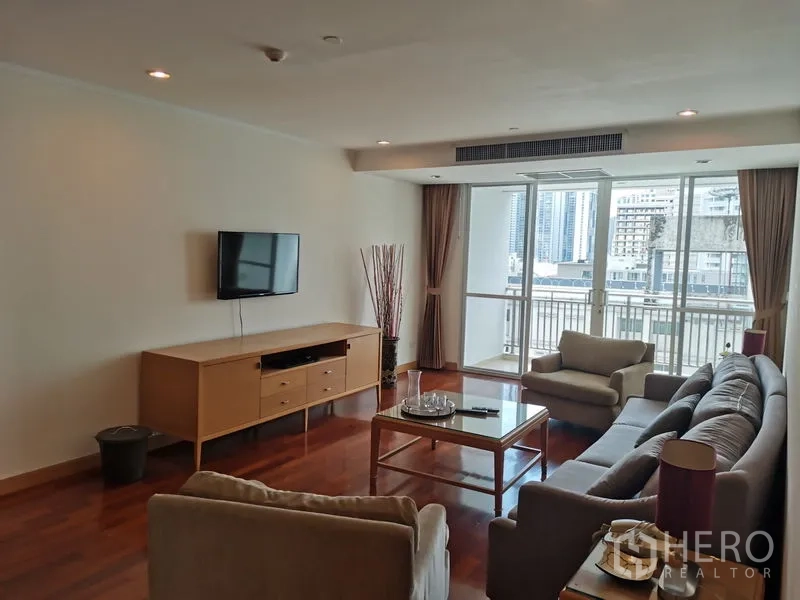 Condominium for rent in Khlong Toei, Bangkok - City-facing living room with sofa set, wall-mounted TV, and large sliding doors to a bright balcony.