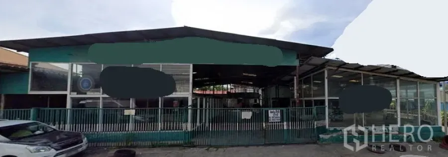 Land for sale in Suan Luang, Bangkok - Street‑facing gated land plot with wide frontage, perimeter fence, metal‑roof shelter and glass‑fronted rooms suitable for storage or workshop.