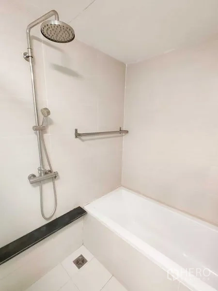 Condominium for rent in Khlong Toei, Bangkok - White bathtub with rainfall showerhead and towel rail in a tiled bathroom.