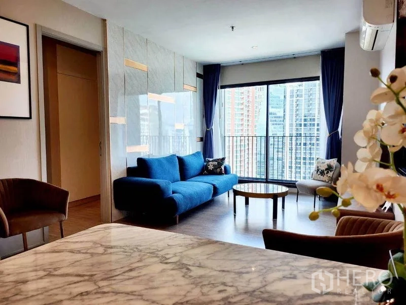 公寓 出租在Watthana, Bangkok - 开放客厅配大理石质感装饰墙、蓝色沙发组，阳台框景高楼林立。