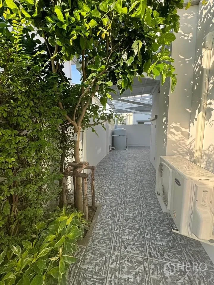 Detached House for rent in Bang Phli, Samut Prakan - Shaded side yard with mature plants, outdoor air-conditioning unit, and a tiled path.