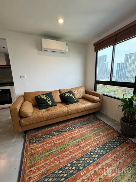 Condominium for rent or sale - Bright living room with tan leather sofa, patterned rug, AC, and skyline view from a wide window.