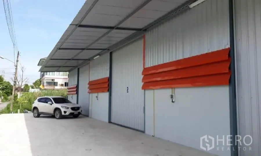 Warehouse for rent in Mueang Pathum Thani, Pathum Thani - Warehouse exterior in Mueang Pathum Thani with wide roller shutter doors, covered loading bay, and parking on a paved driveway.